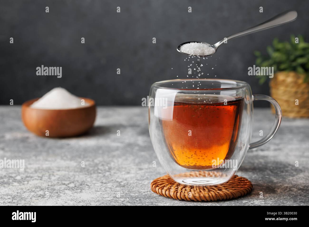 White sugar falling from teaspoon into glass cup of hot tea placed on coaster against grey background Stock Photo