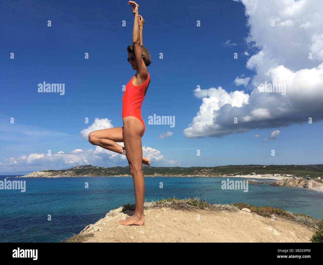 Beautiful sporty woman wearing orange swimsuit doing active pose with one leg up  on the edge of sea view point in Italy in Sardinia. Stock Photo