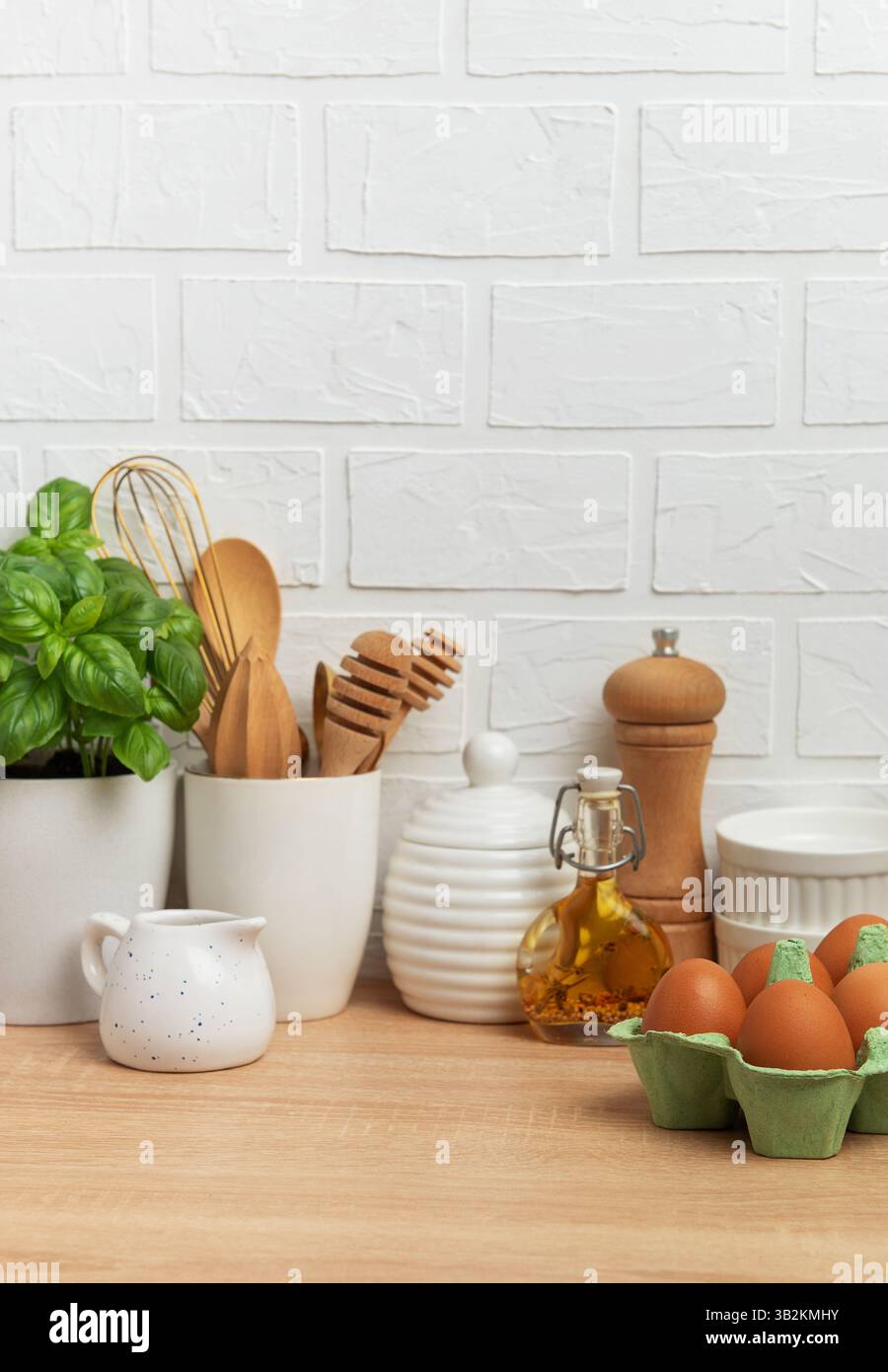 Kitchen counter displaying fresh eggs, basil plant, oil, spices, and various utensils against a white brick wall, embodying a home cooking atmosphere Stock Photo