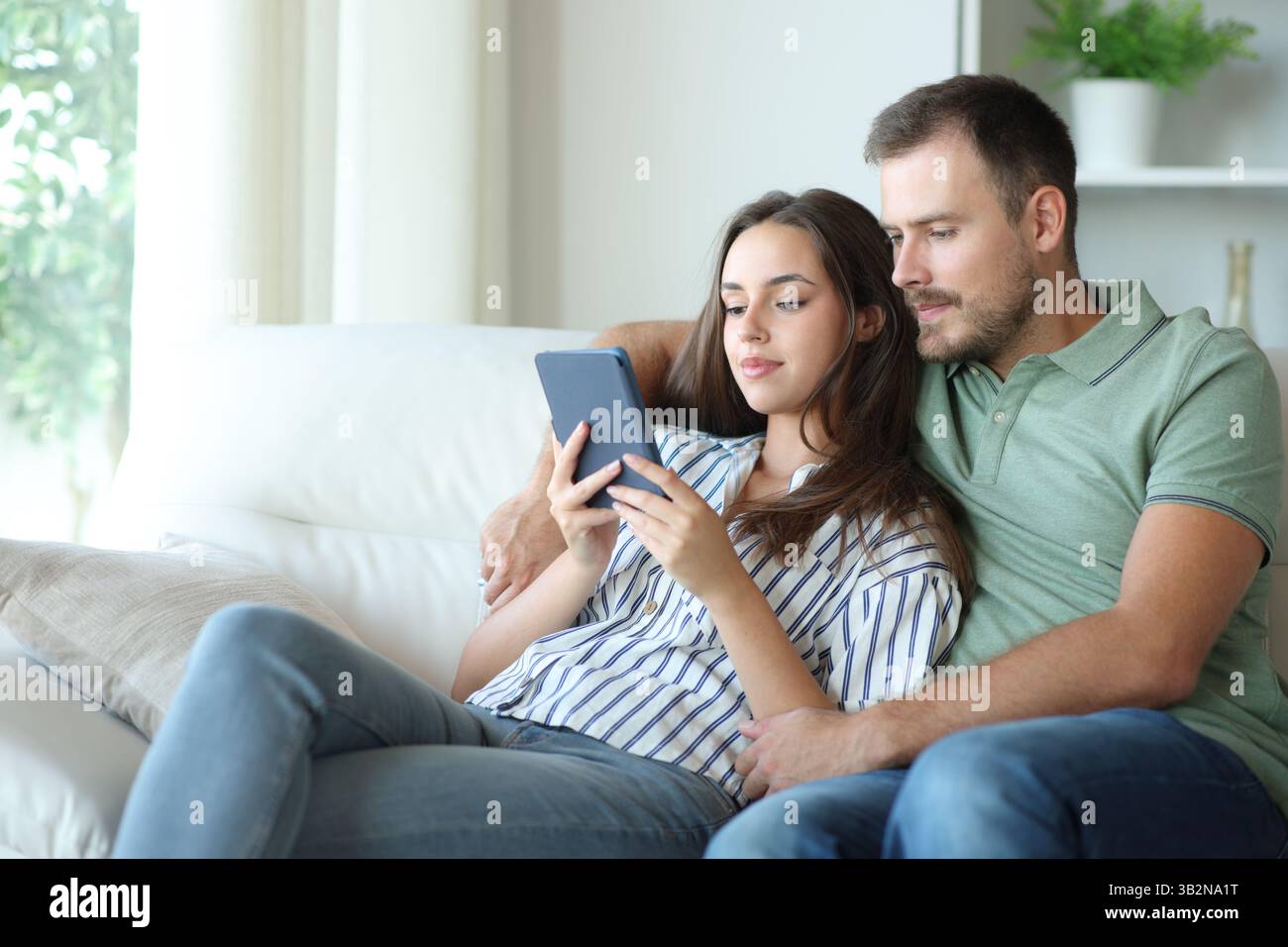 Couple reading together the same ebook sitting on a couch at home Stock Photo