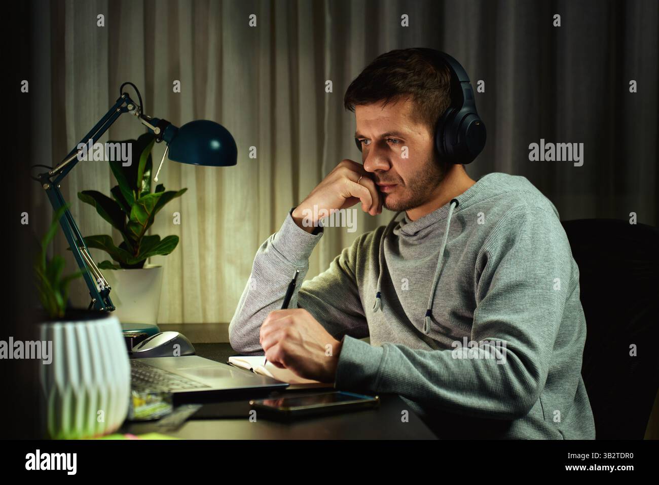 Focused man in headphones working on laptop at night and writing notes. Freelancer working remotely from home Stock Photo