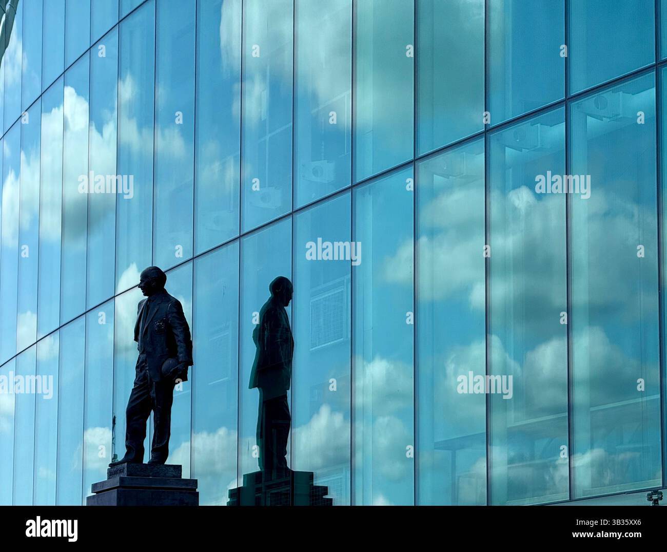 Sir Matt Busby statue at Old Trafford football Stadium. Statue and reflection with clouds and blue sky reflected in windows of stadium front. Manchest - Smartphone Captured Stock Image