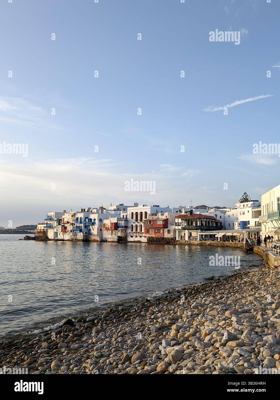 A view of bars and restaurants on the coast of Mykonos Town, Greece, overlooking the Aegean Sea under a blue sky just before the sun sets - Smartphone Captured Stock Image
