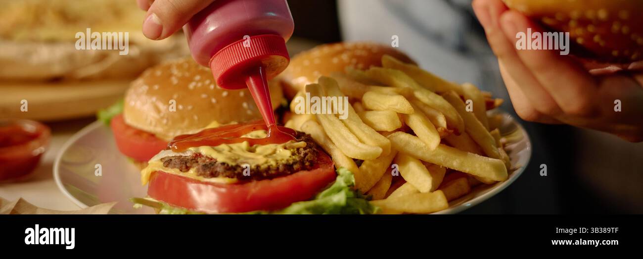 Close-up of human hand adding ketchup onto cheeseburger patty with sesame bun, mustard, and lettuce, with fries in background on plate, depicting casual dining experience Stock Photo