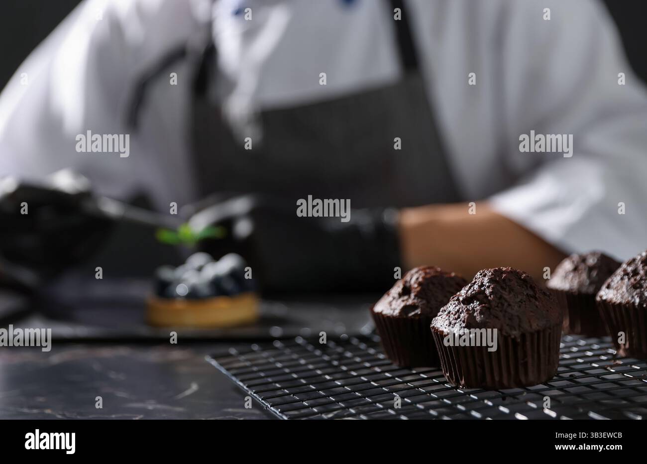 Confectioner decorating tasty tart with mint at dark table, focus on chocolate muffins Stock Photo