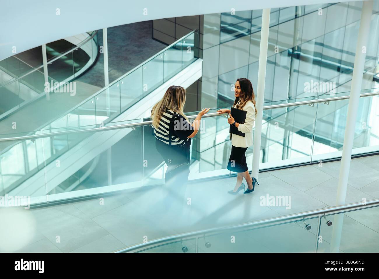 Two female colleagues engage in conversation on a sleek, modern walkway within a corporate office. Dressed in business attire, they appear engaged and Stock Photo