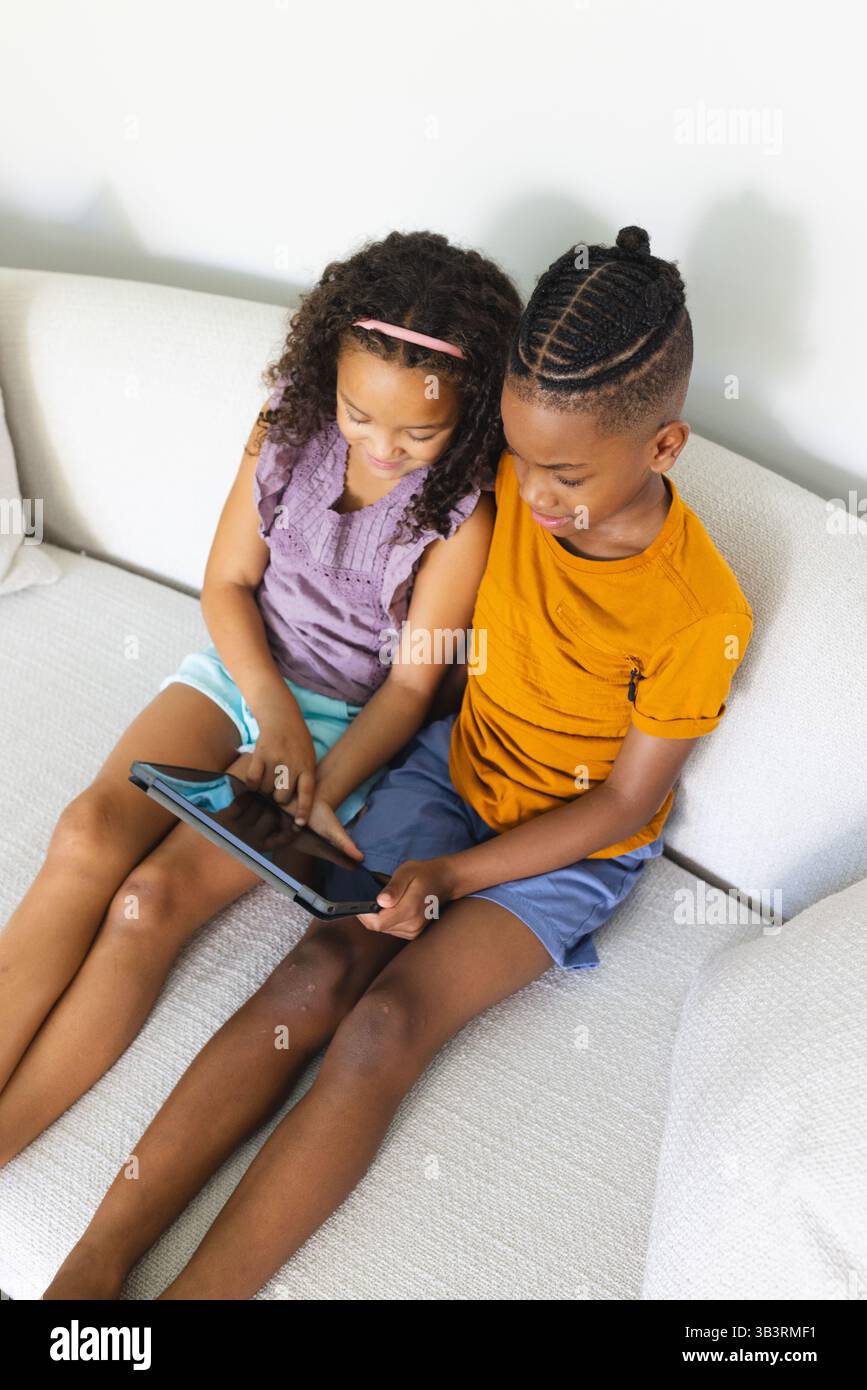 Diverse siblings interacting with black tablet while sitting on light sofa, exploring content Stock Photo