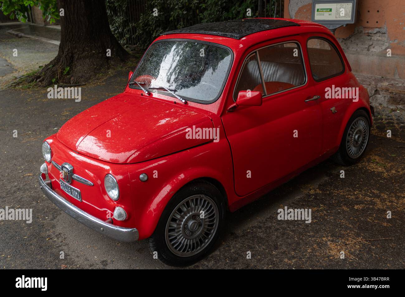 Red Fiat 500, vintage car parked on a street under a tree, featuring an elegant design and raindrops on its surface, Aventino, Rome, Italy Stock Photo
