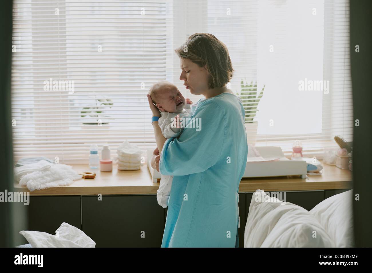 Caucasian woman cradling newborn baby near window in cozy, well-lit room, with baby essentials on the counter, emanating a warm, nurturing atmosphere Stock Photo