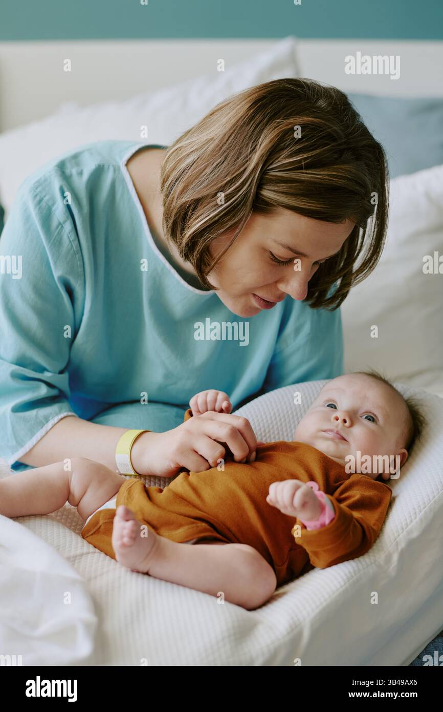 Woman gently embracing her baby while lying on bed, showcasing tender moment filled with affection and love. Infant wearing brown outfit and looking calm Stock Photo