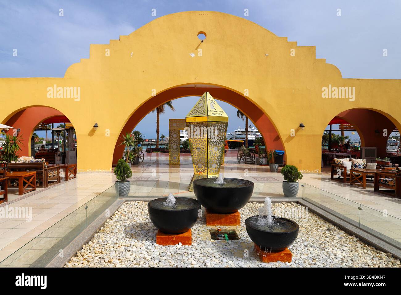 Modern Middle Eastern courtyard with Arabic arches, yellow façade, decorative lantern, and water fountains in Hurghada Marina, Egypt. Stock Photo