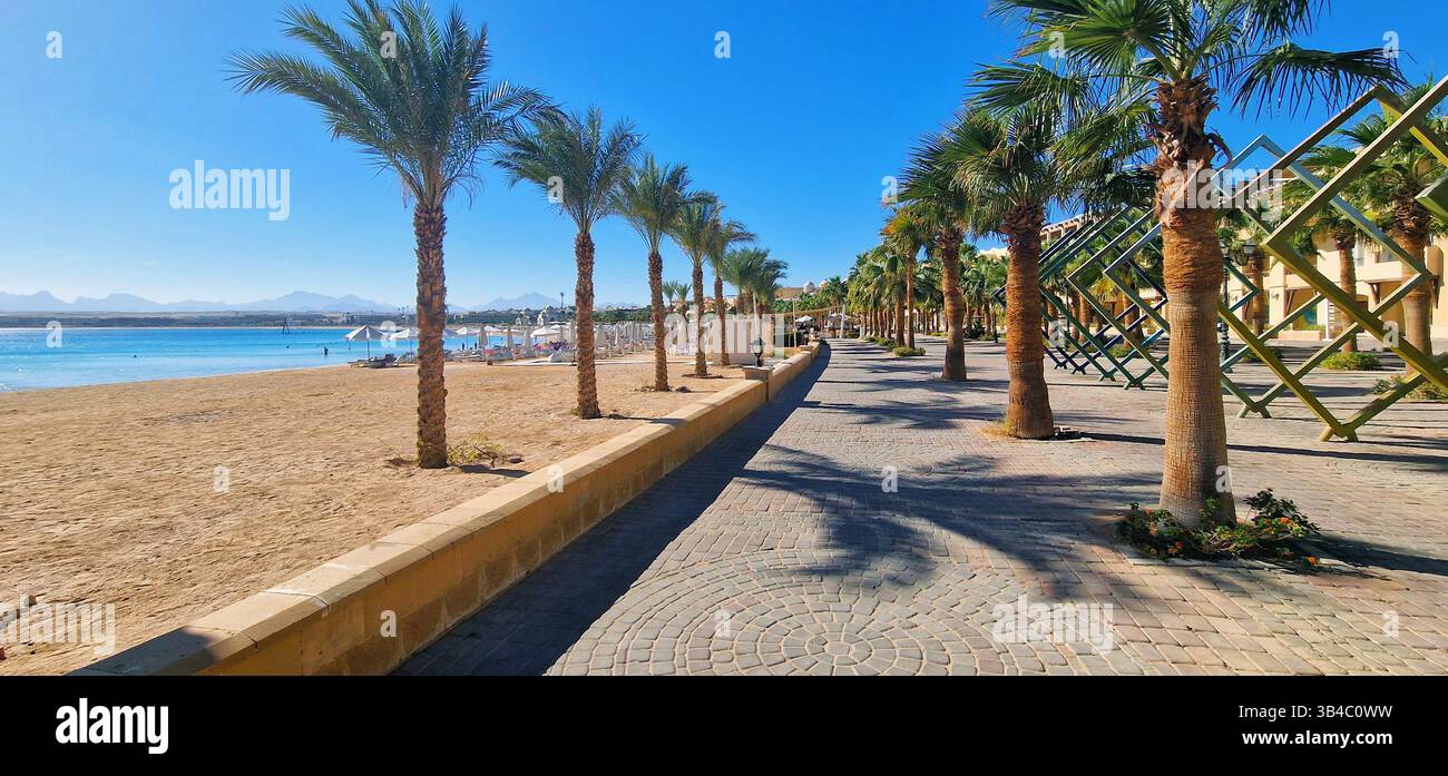 Scenic pedestrian promenade along sandy beach in Sahl Hasheesh, Egypt, with palm trees, beach umbrellas, and a view of the Red Sea coast. Stock Photo