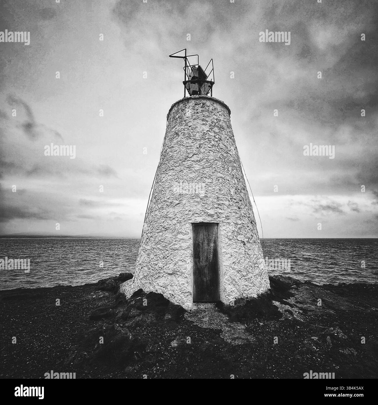 Moody black and white close-up of Ynys Llanddwyn lighthouse, Anglesey, with sea, sky, and coastal terrain in background. - Smartphone Captured Stock Image