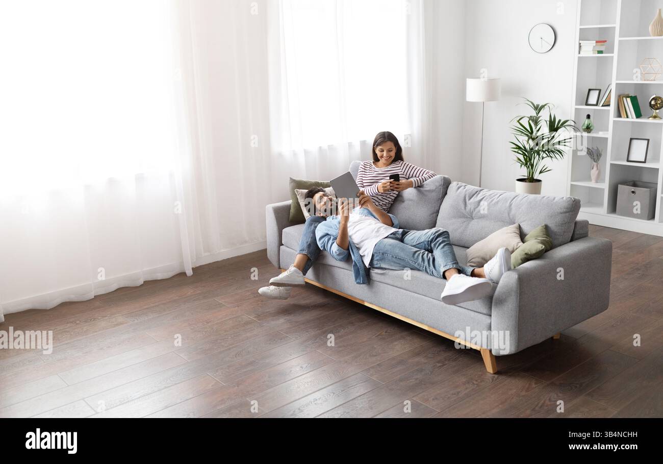 Mixed race spouses resting on sofa at home, using gadgets Stock Photo