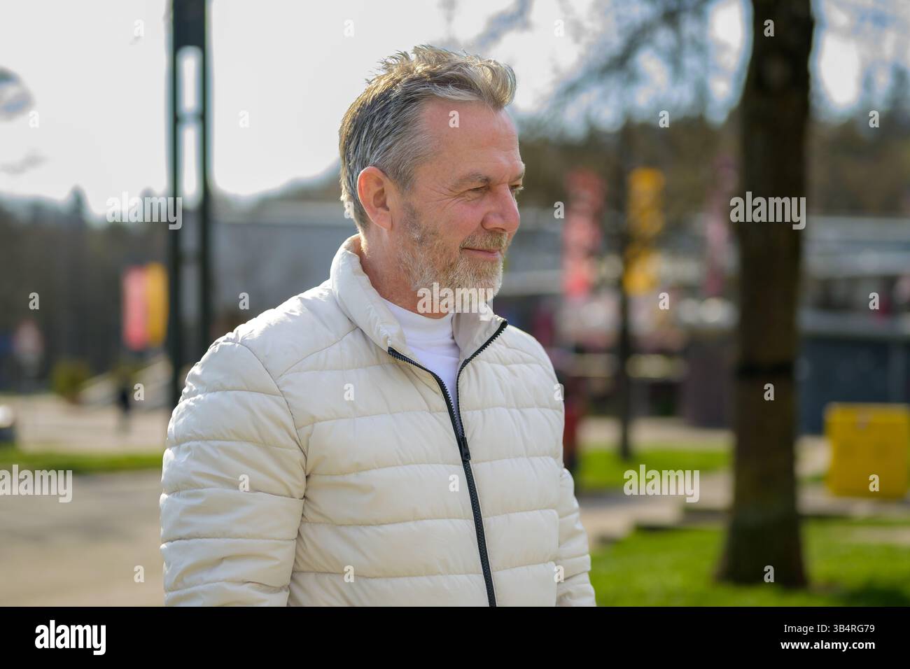 A thoughtful older man stands in a sunlit park wearing a light beige puffer jacket. Stock Photo