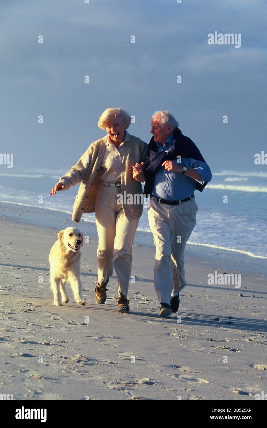 Senior man & woman running on the beach with their dog. Stock Photo