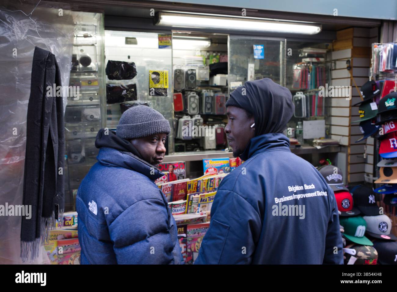 Two men in winter jackets converse outside a newsstand stocked with magazines, phone cases, and hats.  One man wears a dark navy jacket with a logo. T Stock Photo