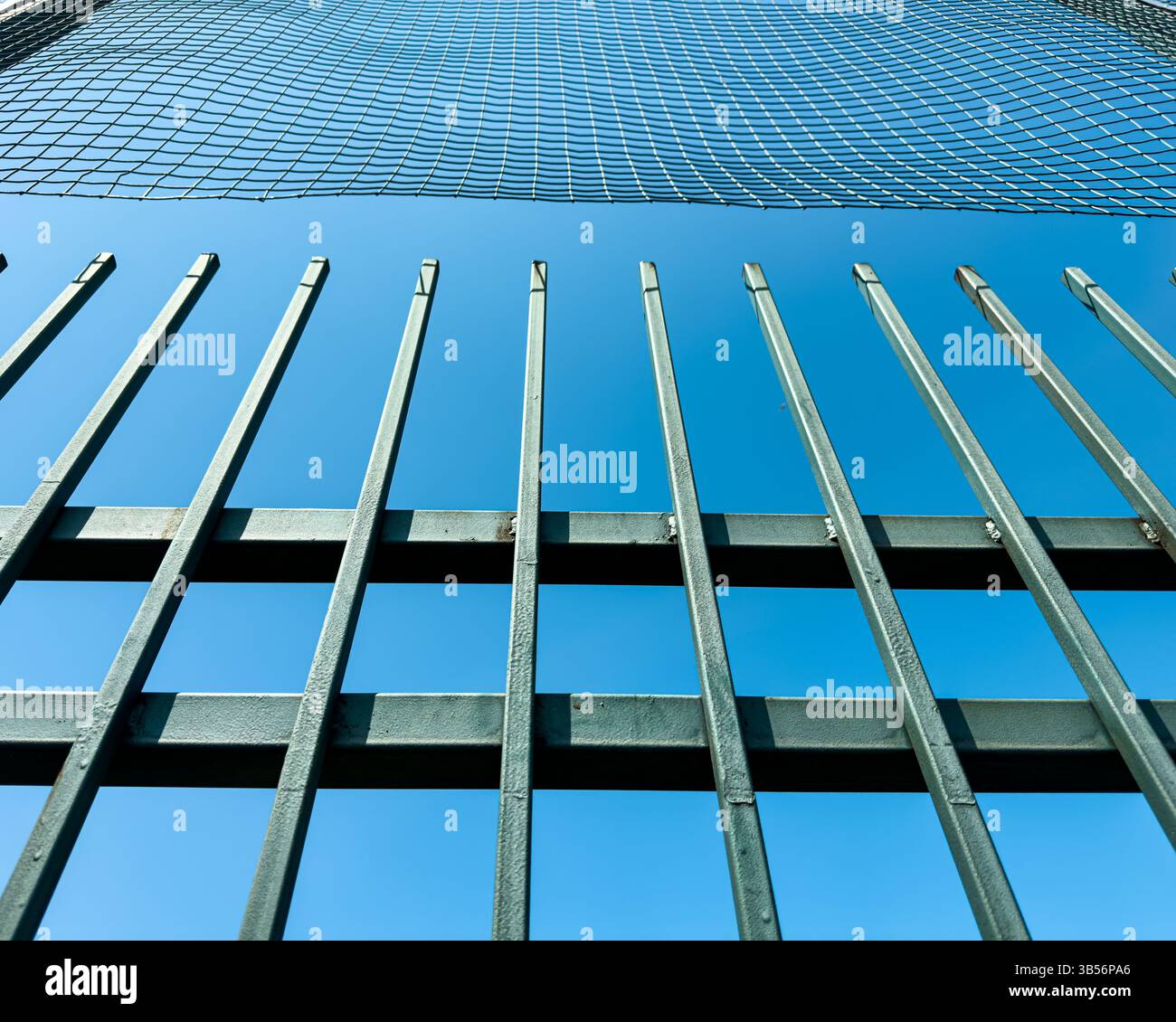 Metal fence with vertical bars and protective netting against clear blue sky. Urban detail, geometric structure, no people, abstract composition. - Smartphone Captured Stock Image