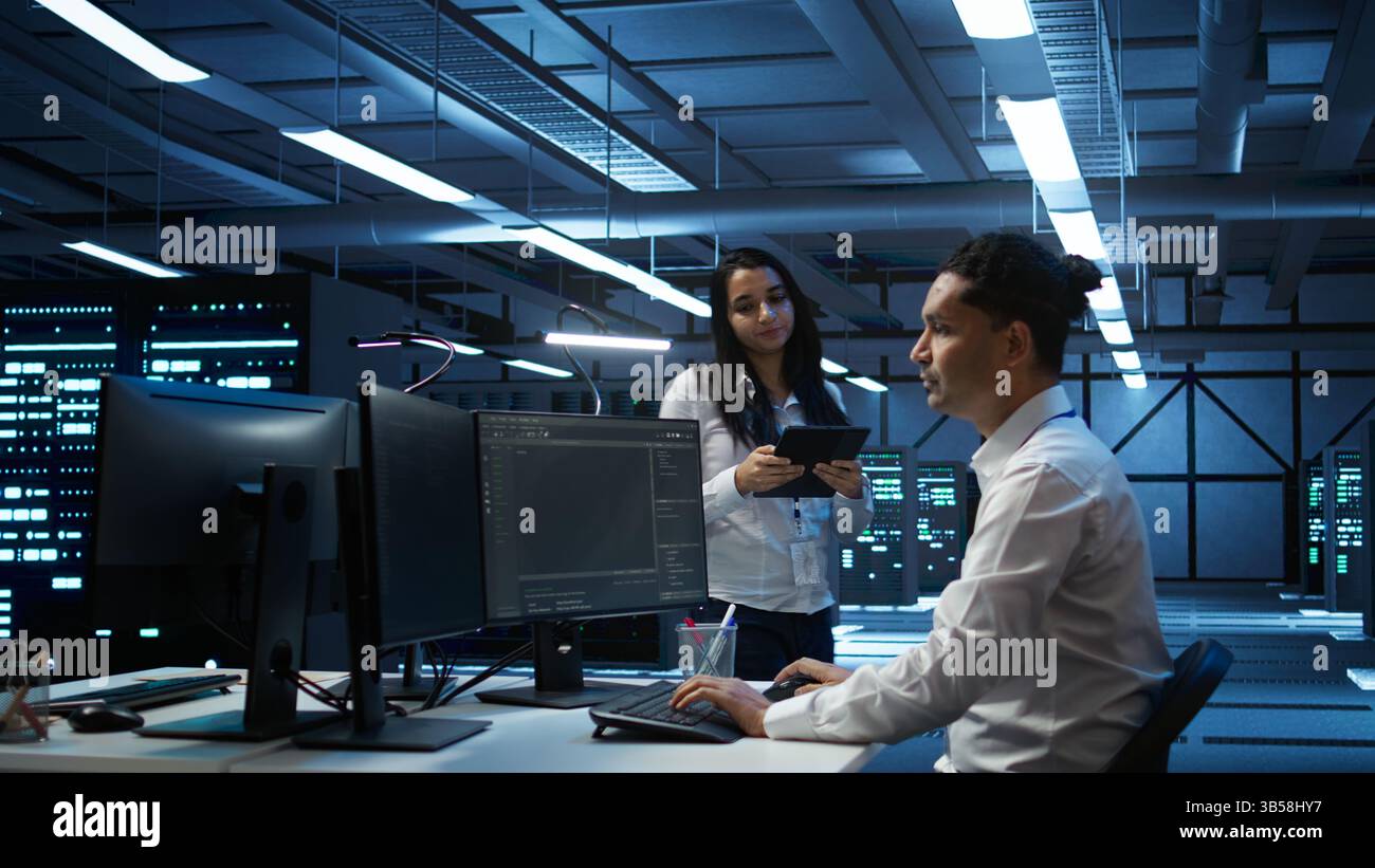 Team of programmers in high tech data center troubleshooting issues using tablet. Teamworking colleagues in server farm using device, establishing performance benchmarks, camera B Stock Photo