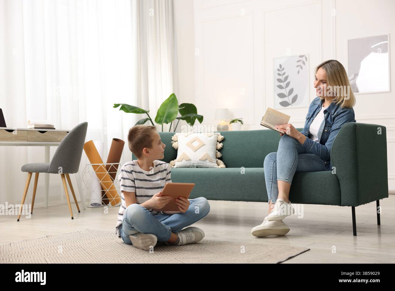 Son using tablet while his mother reading book at home Stock Photo