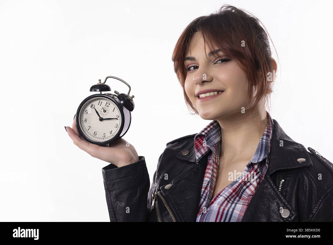 A cheerful young woman confidently poses with an alarm clock, showcasing her style and time management skills Stock Photo