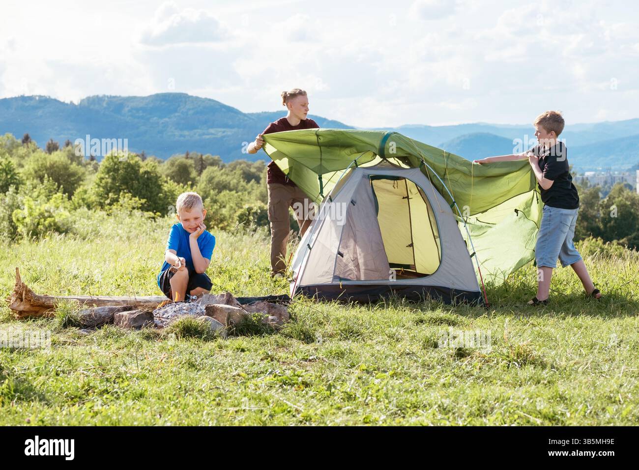 Three brothers have picnic. Boy roasting sausages on sticks over campfire flame while two brothers set up the green tent. Happy family outdoor picnic Stock Photo