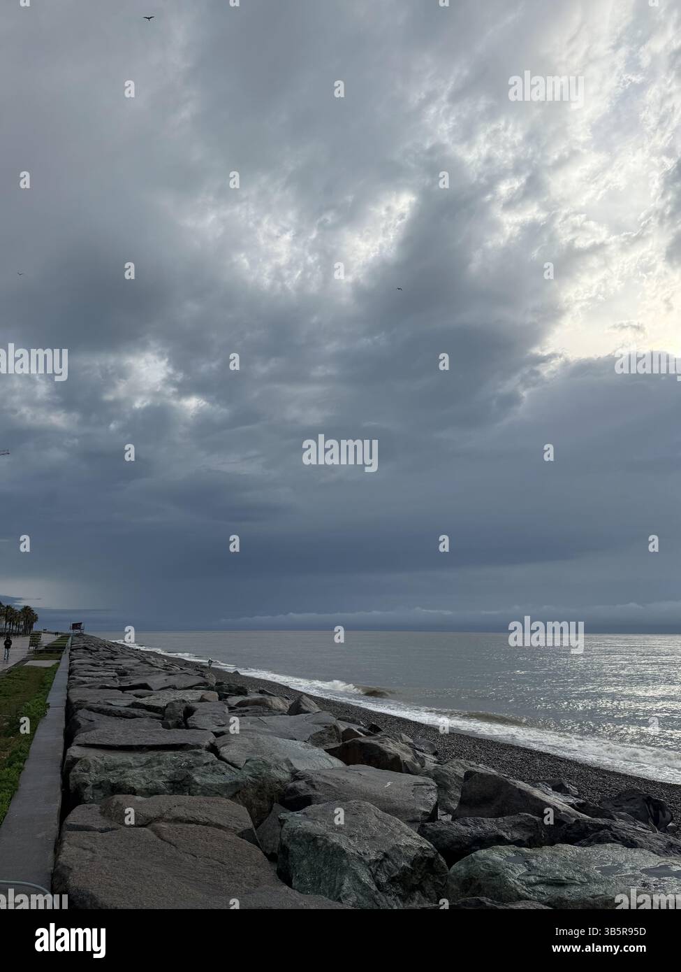The image shows a rocky shoreline beside a pebble beach with waves gently breaking under a cloudy, overcast sky. The scene is framed by large rocks an Stock Photo