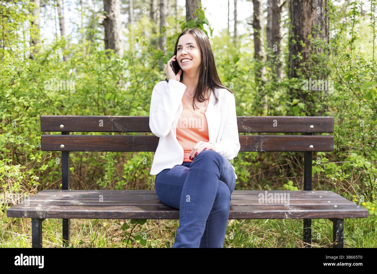 Attractive smiling young woman dressed casually talks with her smartphone sitting on a wooden bench in the forest. Modern lifestyle using portable mob Stock Photo