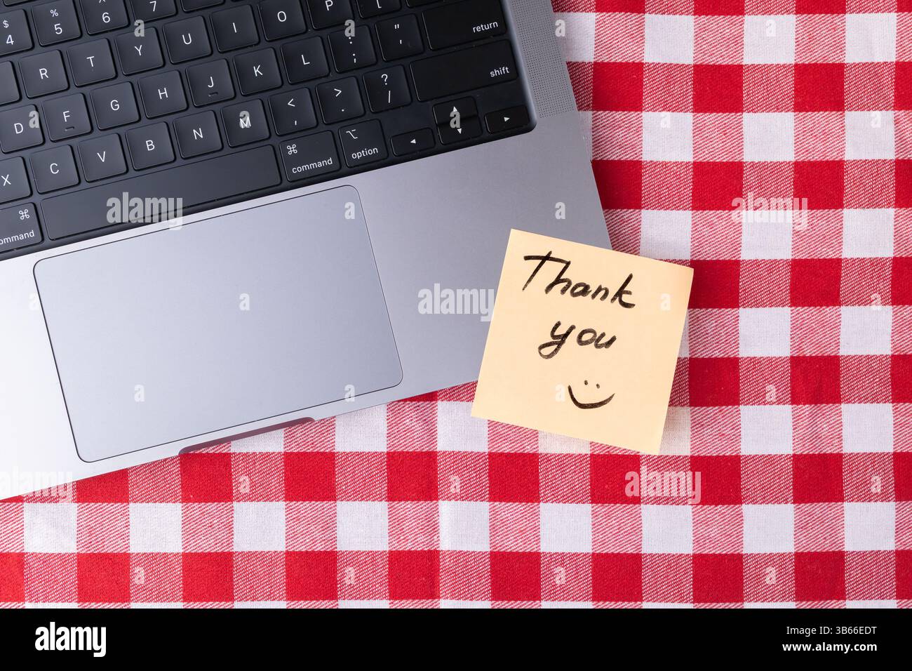 Simple remote workspace featuring a laptop and a bright yellow Thank you note on a red and white checkered tablecloth, perfect for conveying gratitude Stock Photo