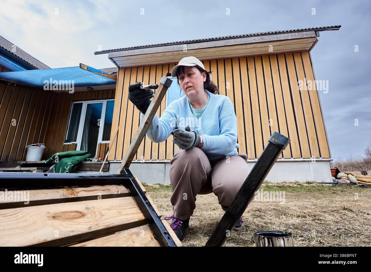 Brushing protective paint onto metal legs of garden bench, dedicated outdoor renovator works methodically, intent on sealing each joint and surface. Stock Photo