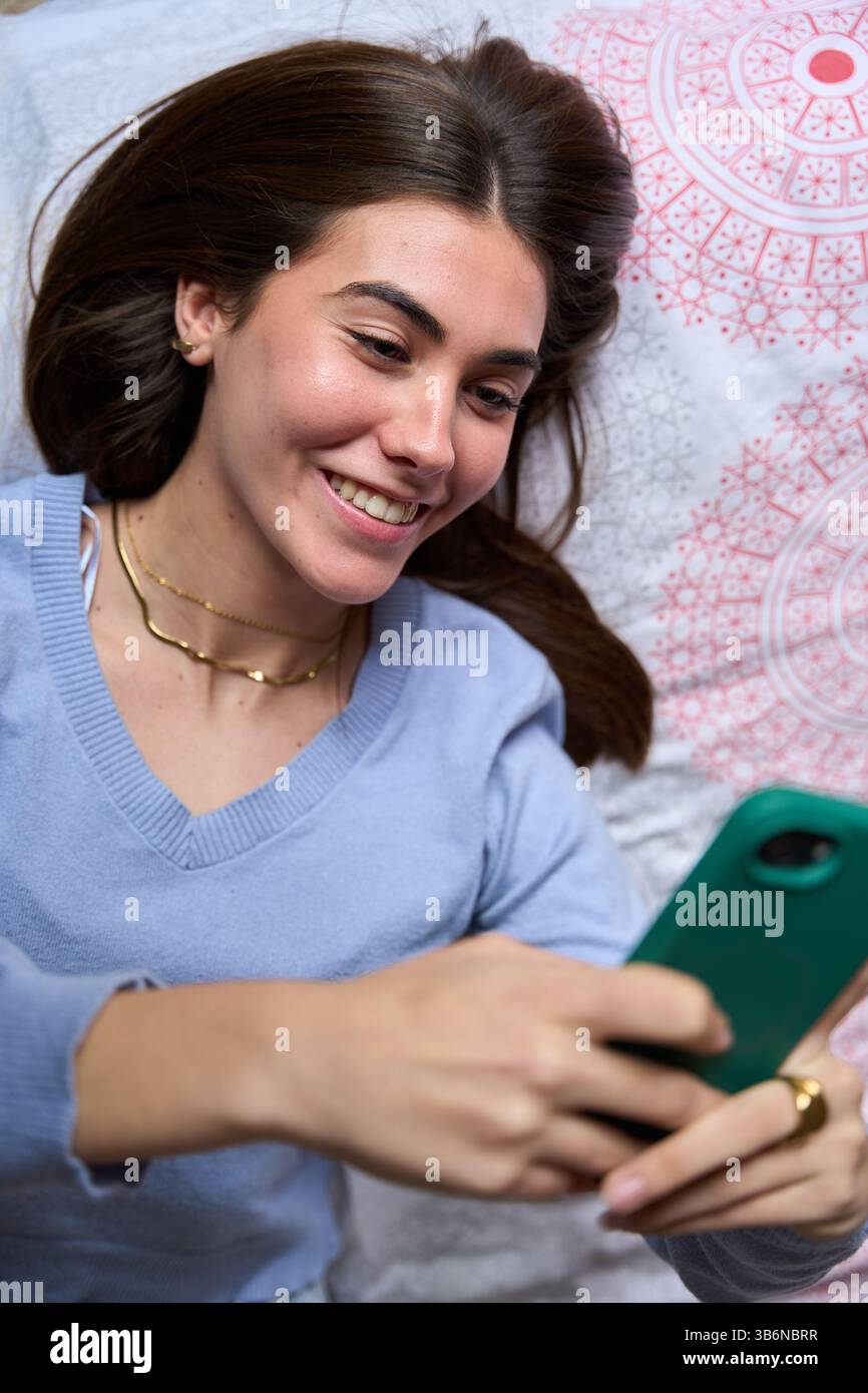 Happy female student using smartphone lying on bed Stock Photo