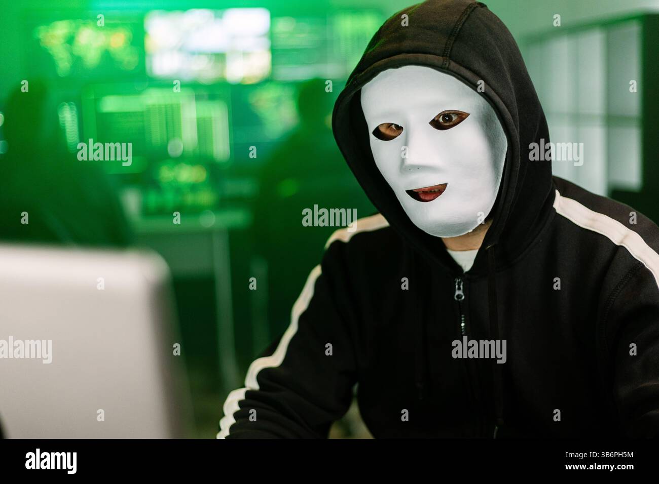 Hacker working on laptop in dark room with green code displayed on monitors Stock Photo