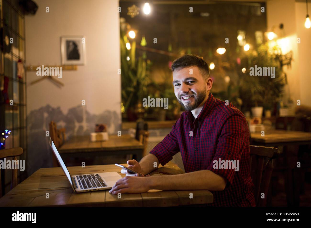 A young handsome Caucasian man with beard and toothy smile in red checkered shirt is working behind laptop sitting at wooden table. Uses holds mobile Stock Photo