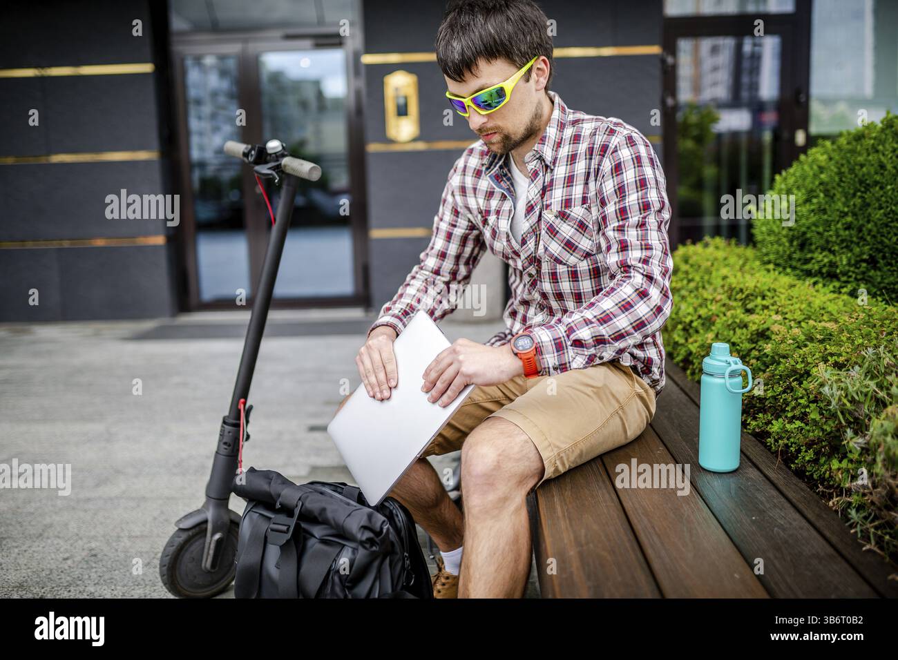 Young caucasian man in casual clothes freelancer uses eco-friendly electric transport scooter. Male takes out laptop from backpack, sitting on bench n Stock Photo