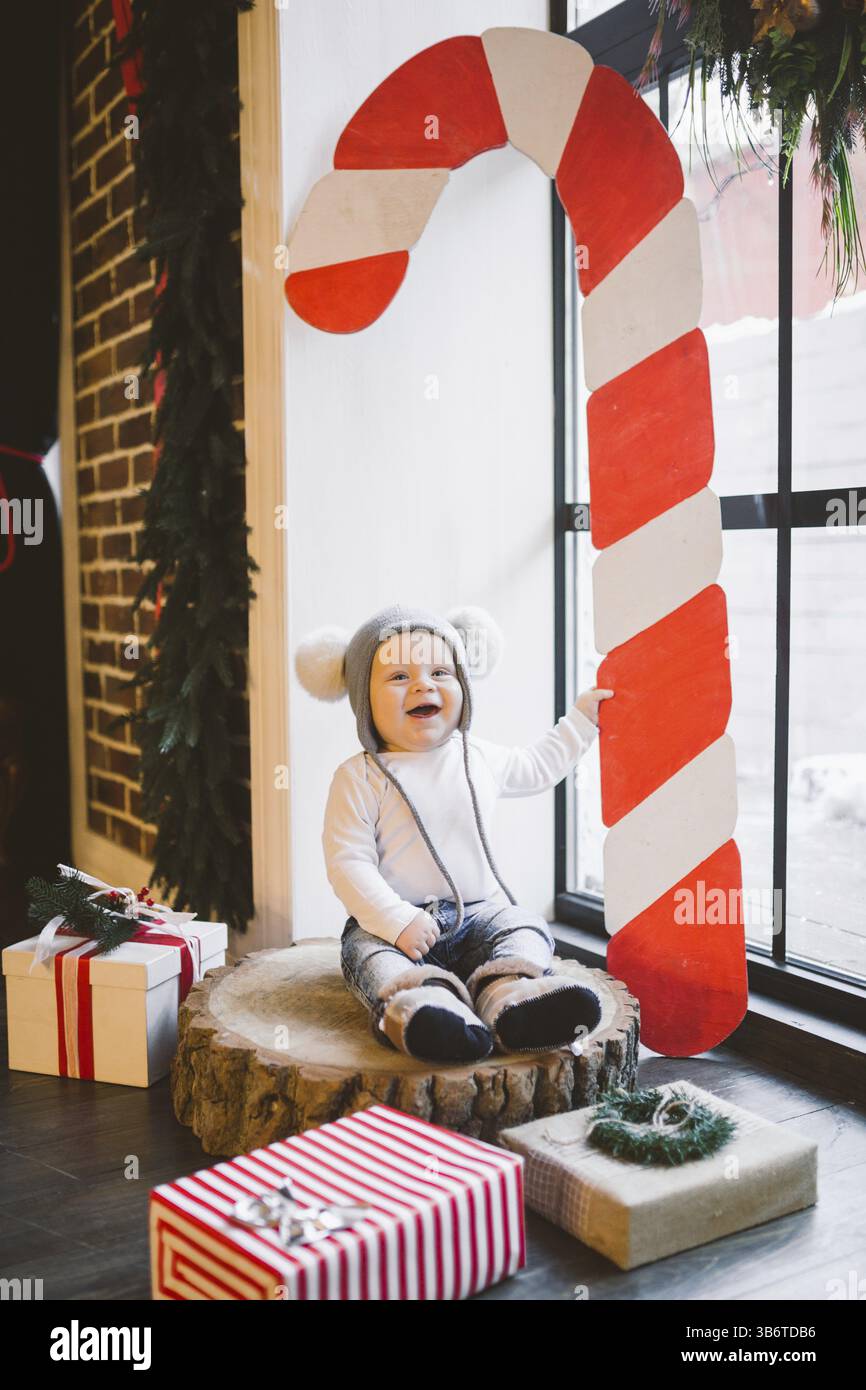 New year and Christmas holidays theme Caucasian child boy 1 year old sitting on a stump felled tree near the window in a funny hat. Scenery large red Stock Photo