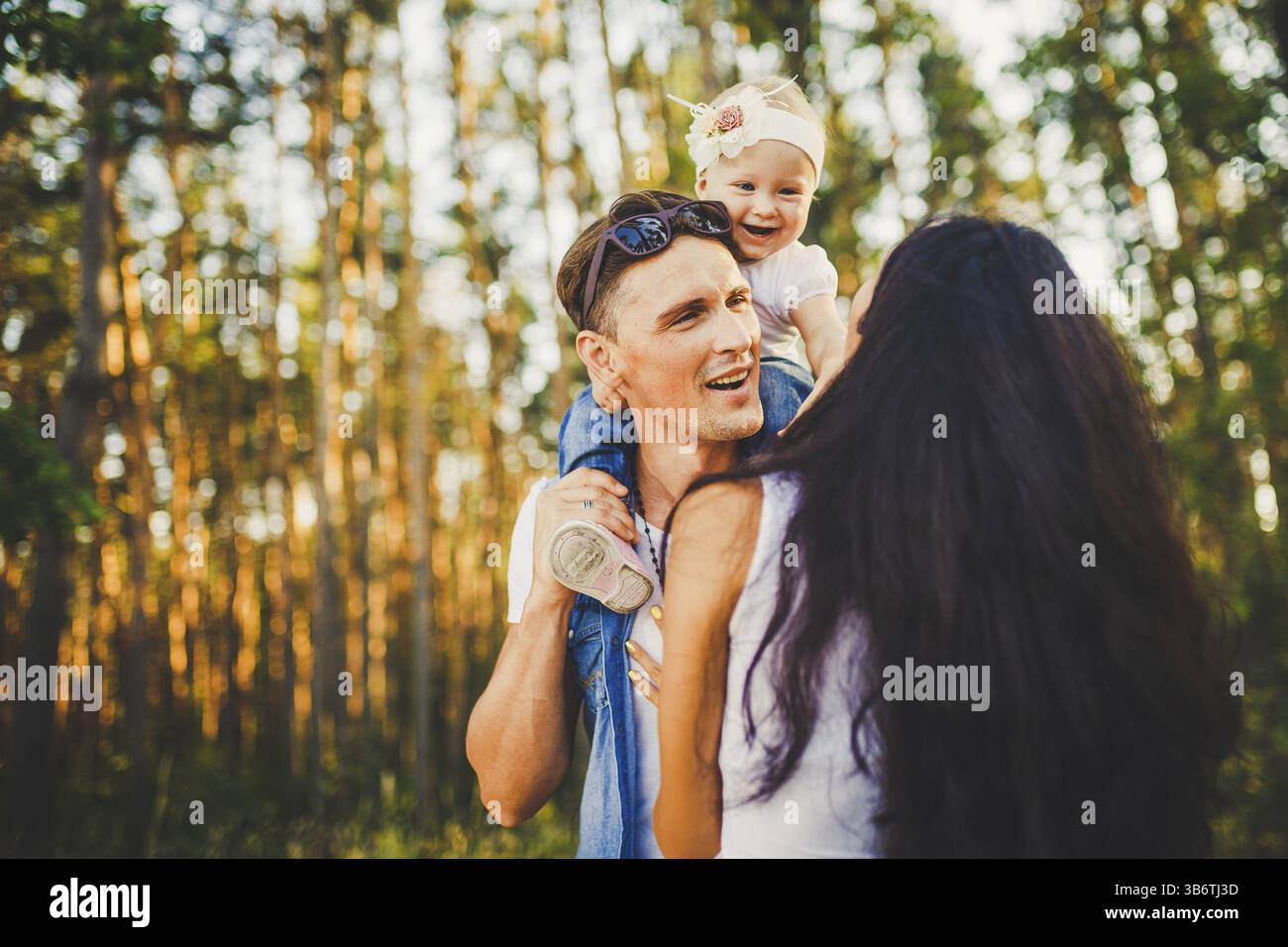 Stylish young Family of mom, dad and daughter one year old blonde sitting near father on shoulders, outdoors outside the city in a park amid trees in Stock Photo