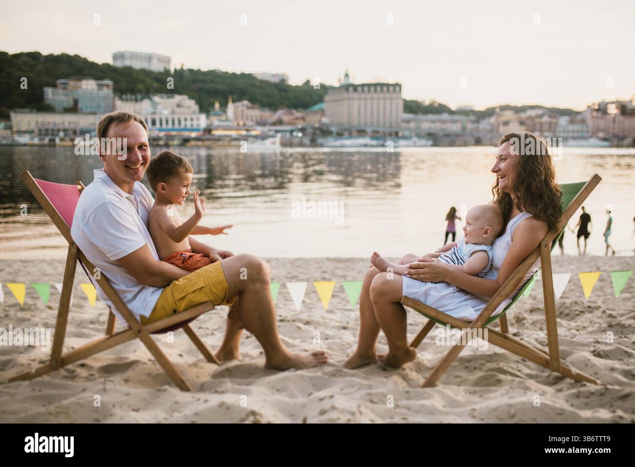 Family at seaside in evening open-air cafe. Mother and father and two sons sit on sun loungers, looking at sunset on sandy beach near river overlookin Stock Photo