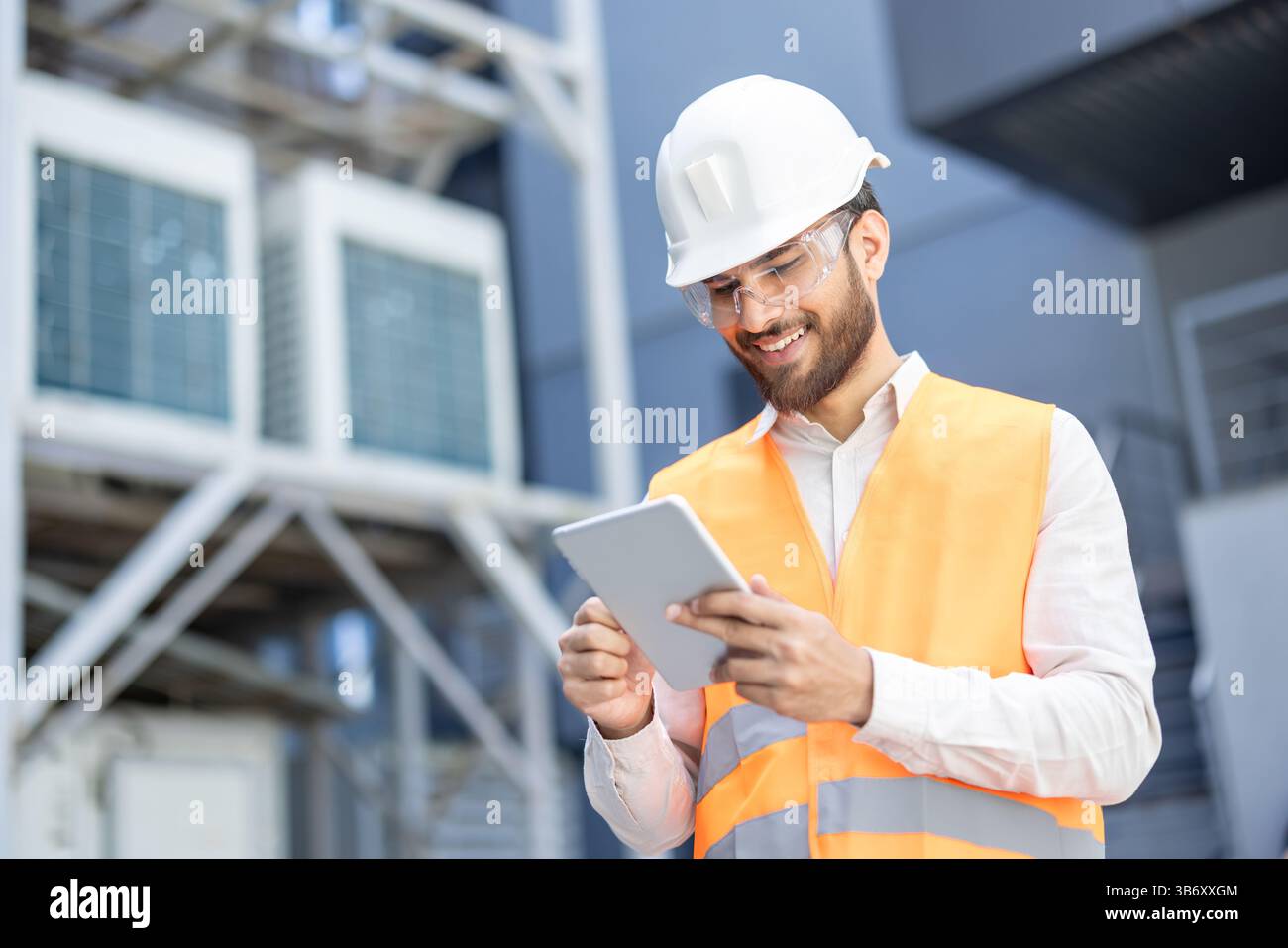 A smiling engineer in a white helmet and safety vest uses a tablet in a industrial setting, possibly for equipment inspections. Stock Photo