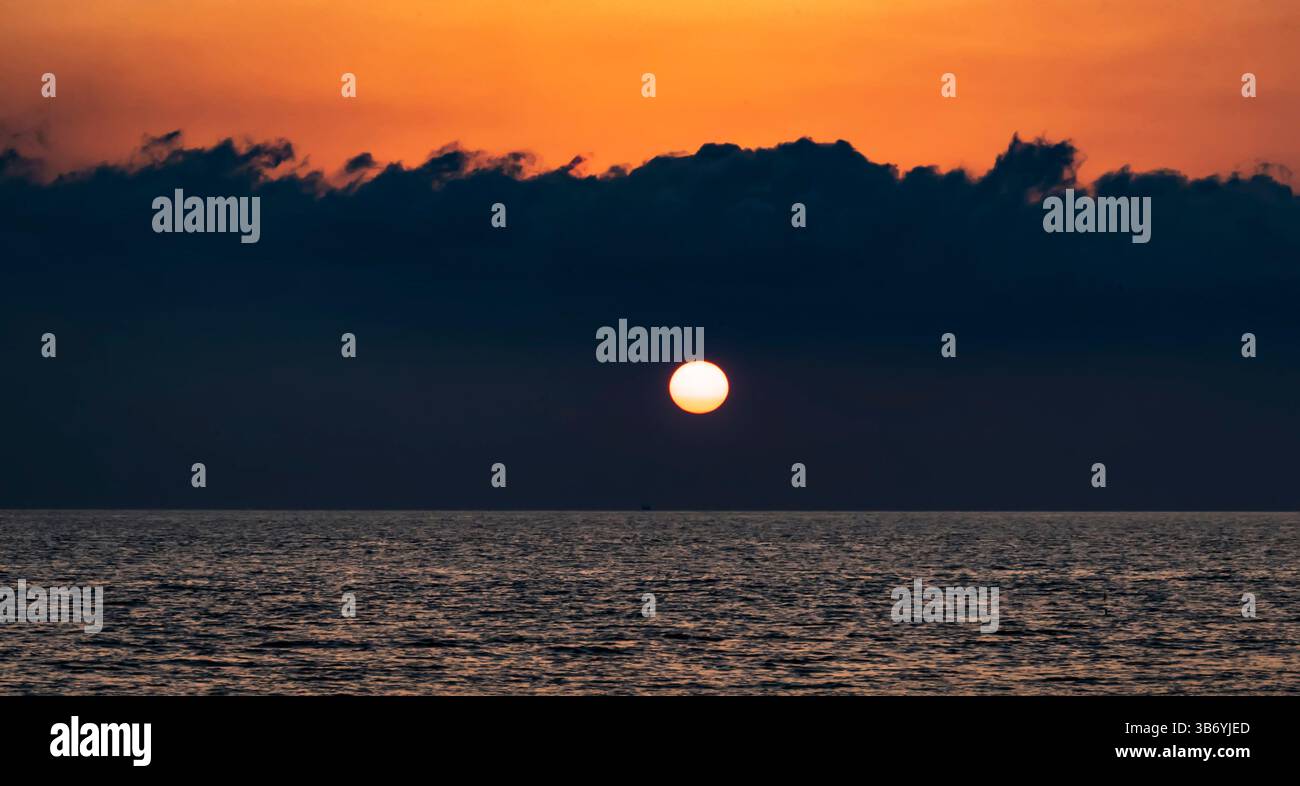 A serene sunset over the ocean, with a vibrant orange sky and dark clouds. The sun is setting on the horizon, casting a warm glow on the water's surfa Stock Photo