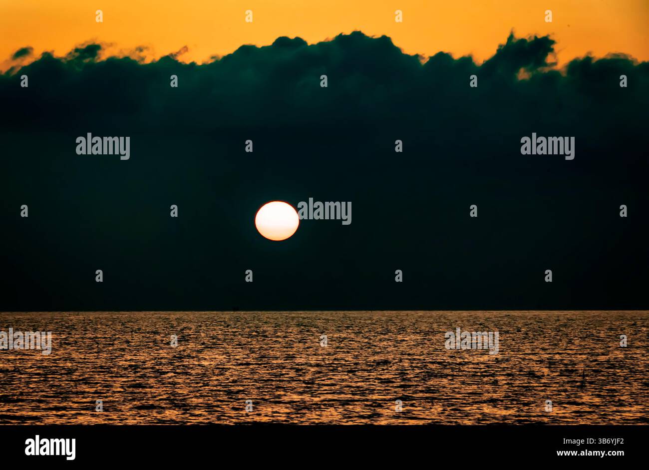 A serene sunset over the ocean, with a large sun setting on the horizon. The sky is a gradient of orange and yellow, contrasted by dark clouds. The wa Stock Photo