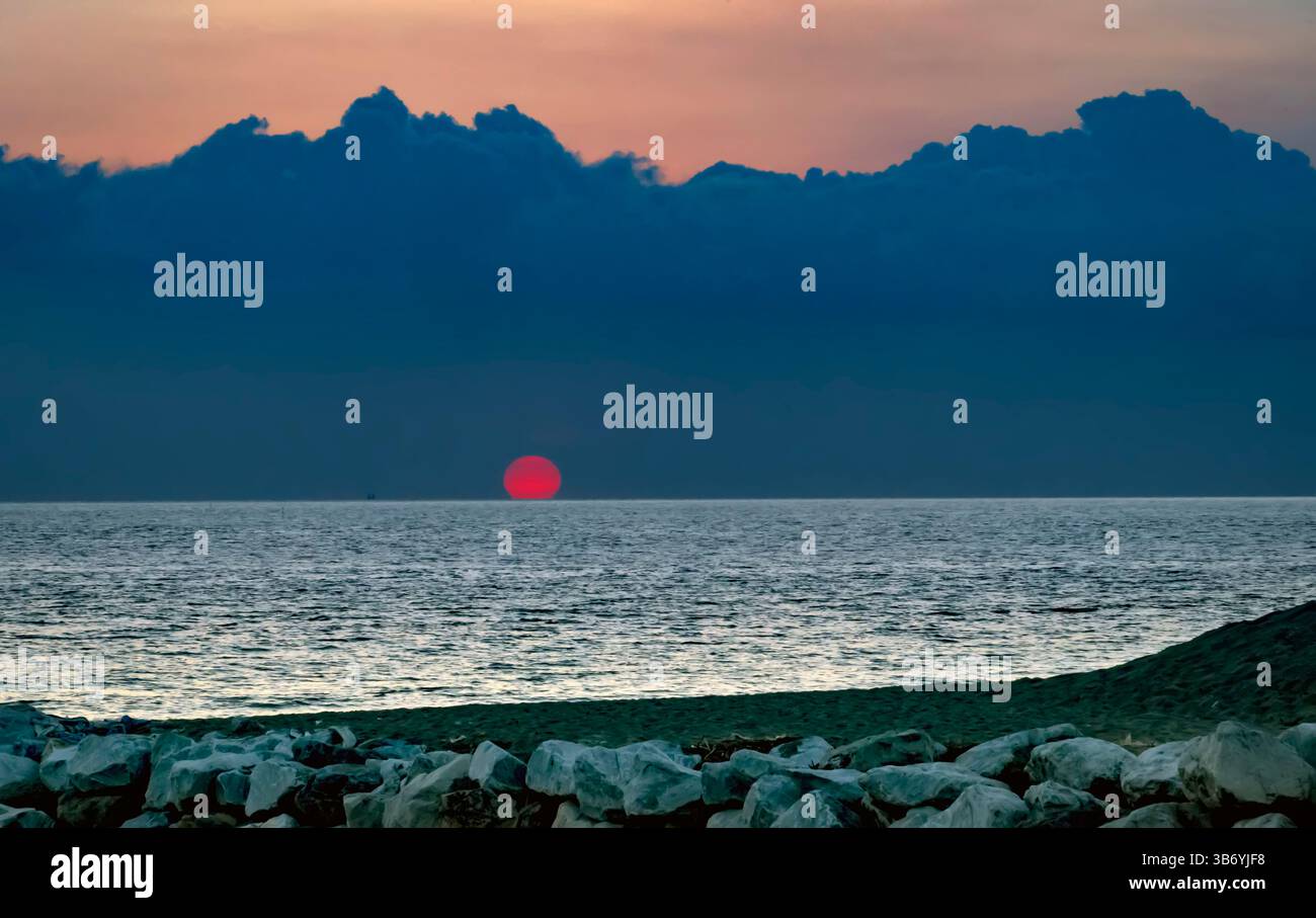 A serene sunset over the ocean, with the sun partially hidden behind clouds. The sky is painted in hues of orange and blue, reflecting on the calm wat Stock Photo