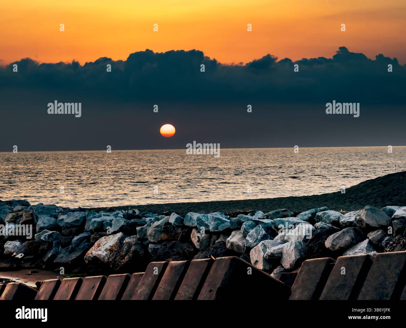 A serene sunset over the ocean, with the sun partially hidden behind dark clouds. The foreground features a rocky shoreline and wooden structures, cre Stock Photo