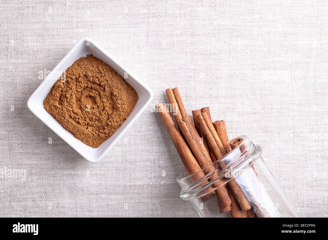 Cassia cinnamon powder in a white bowl, with cinnamon sticks sticking out of an open screw-top jar, on linen fabric, with empty space for text. Stock Photo