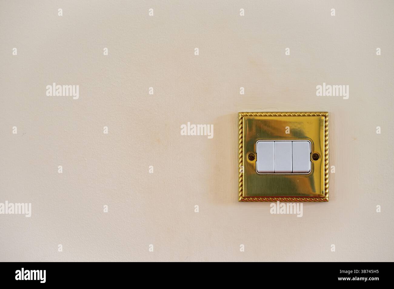 Triple light switch. Gold switch with white details and magnolia wall. All switches in off position. No branding. Copy space to left. Stock Photo