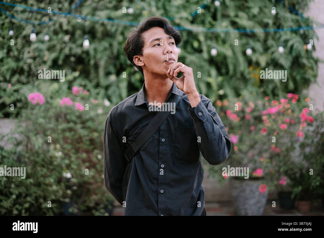 Young man holding a cigarette, taking a contemplative moment amidst lush greenery outdoors. Casual style with a thoughtful expression Stock Photo
