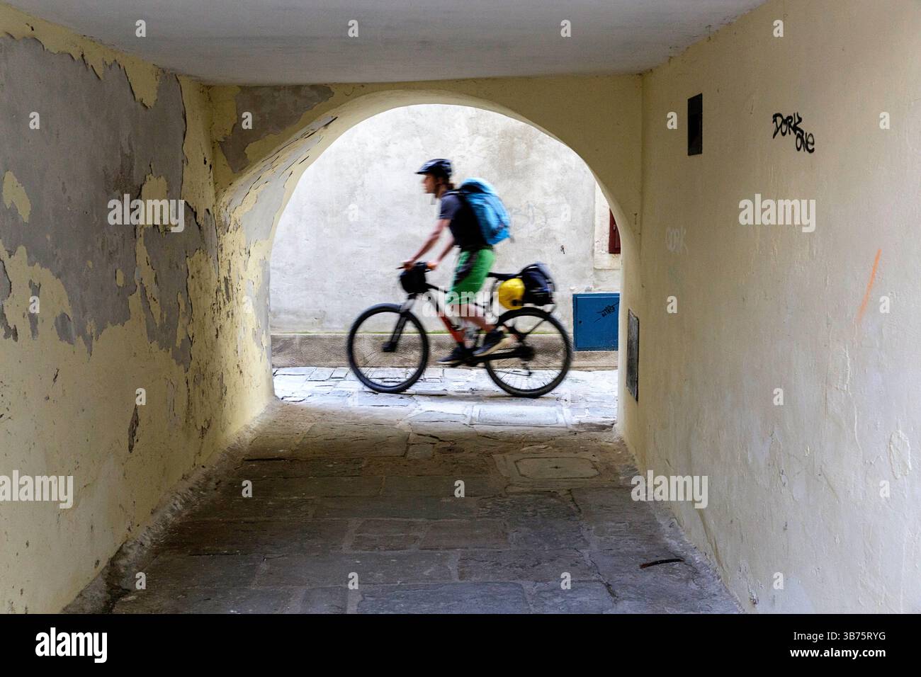 Tourist on a cycling, bikepacking trip from Ljubljana to Piran in Slovenia, cycling in old narrow cobbled streets of piran town, passing stone arch Stock Photo