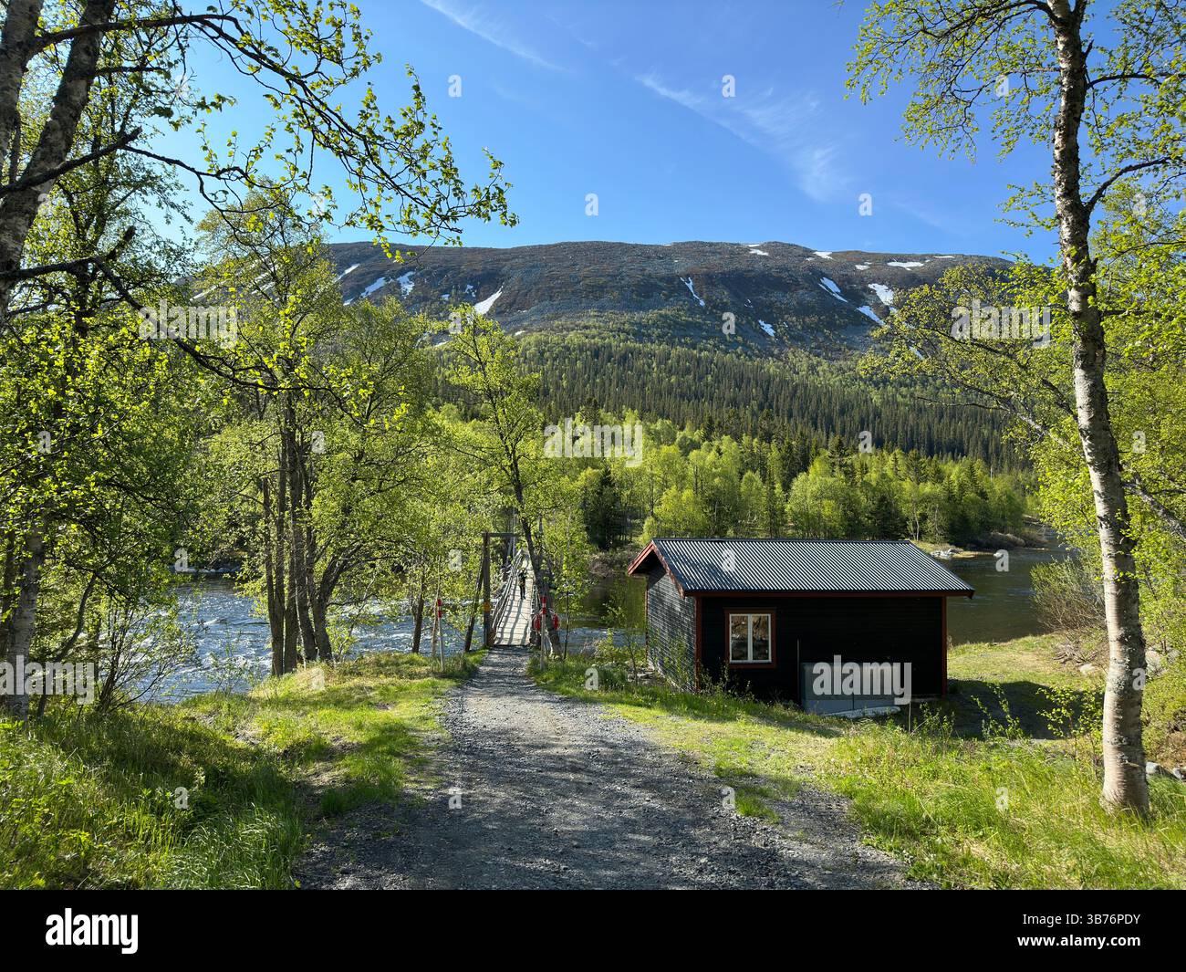 A cabin nestled near a river with a suspension bridge and a mountain backdrop in north Sweden. Stock Photo