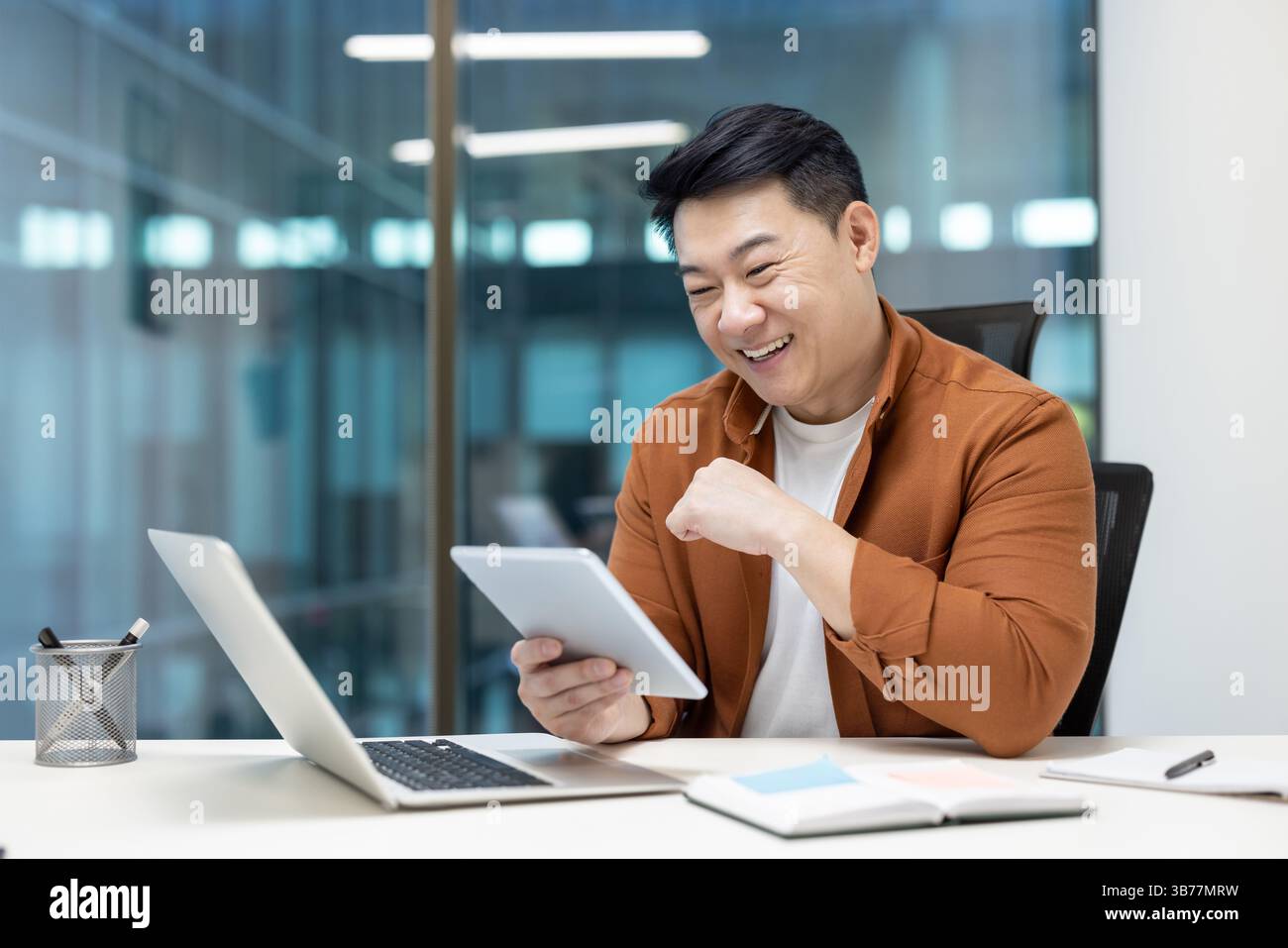 A cheerful programmer enjoys testing an application on a tablet, seated at a modern office desk with a laptop Stock Photo