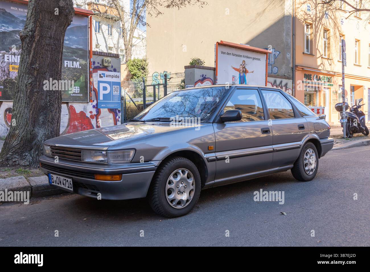 Classic silver Toyota Corolla sedan parked on a city street in front of colorful urban walls and posters. A nostalgic scene of everyday life in a Euro Stock Photo