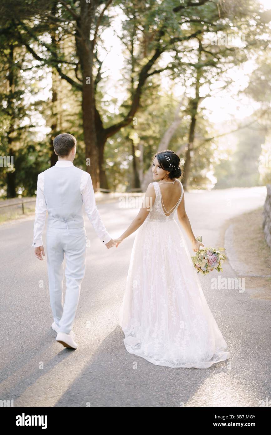The bride and walk holding hands on the road among the trees in an ...